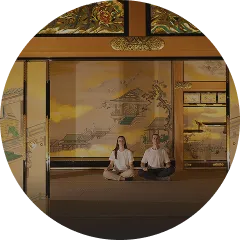 山水画が描かれた黄金色の障壁の前で、白いシャツ姿の二人が床に座り瞑想しています。