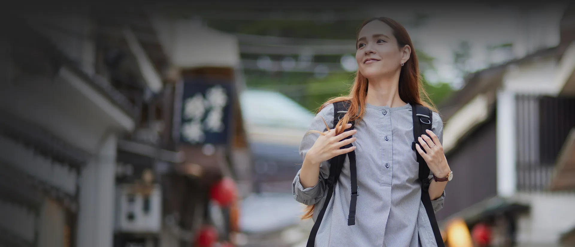 バックパックを背負った女性が、伝統的な日本の街並みの中で笑顔を浮かべ、遠くを眺めている様子。