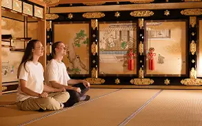 豪華な金屏風や障子がある和室の畳の上で、2人の人物が並んで静かに座禅（瞑想）をしている様子。