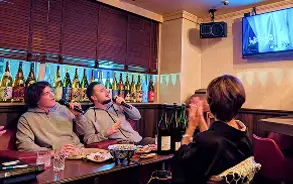 お酒のボトルが並ぶ飲食店で、数人のグループがテーブルを囲み、賑やかに乾杯をしたり会話を楽しんだりしている様子。
