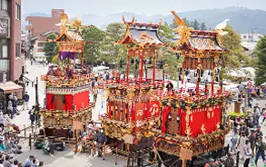 晴天の下、多くの人々が集まる屋外の広場や通りに、豪華絢爛な2台の大きな祭り屋台（山車）が並んで展示されている様子。