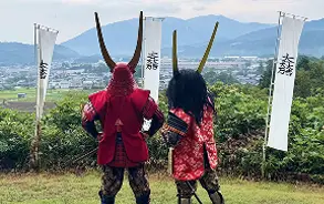 甲冑や大きな角のような兜を身につけた2人の武者姿の人物が、のぼり旗が立つ緑豊かな丘の上から遠くの街並みを眺めている後ろ姿。