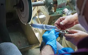 青いゴム手袋をはめた職人の手が、回転する研磨機（グラインダー）を使い、刃物（包丁など）を丁寧に研いでいる様子。