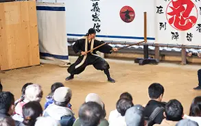 多くの観客が見守る前で、黒い衣装を身にまとった忍者が刀を構えて演技を披露しているステージの様子。
