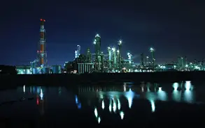夜の闇の中にライトアップされた大規模な化学工場やコンビナートの夜景と、それが静かな水面に美しく反射している様子。