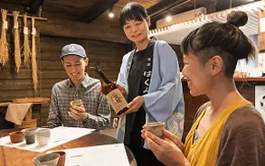 木造の落ち着いたカウンター席で、女性スタッフからお酒のボトルの説明を受けながら、笑顔で試飲を楽しんでいる2人の女性。